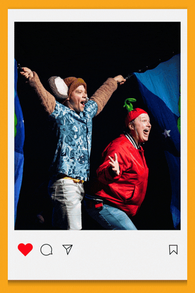 Square Instagram-style image of two performers mid-performance on a stage with a starry blue backdrop, arms raised. One wears a knitted monkey hat and patterned shirt; the other wears a red jacket with a green pom-pom hat, both mid-laugh or mid-song.