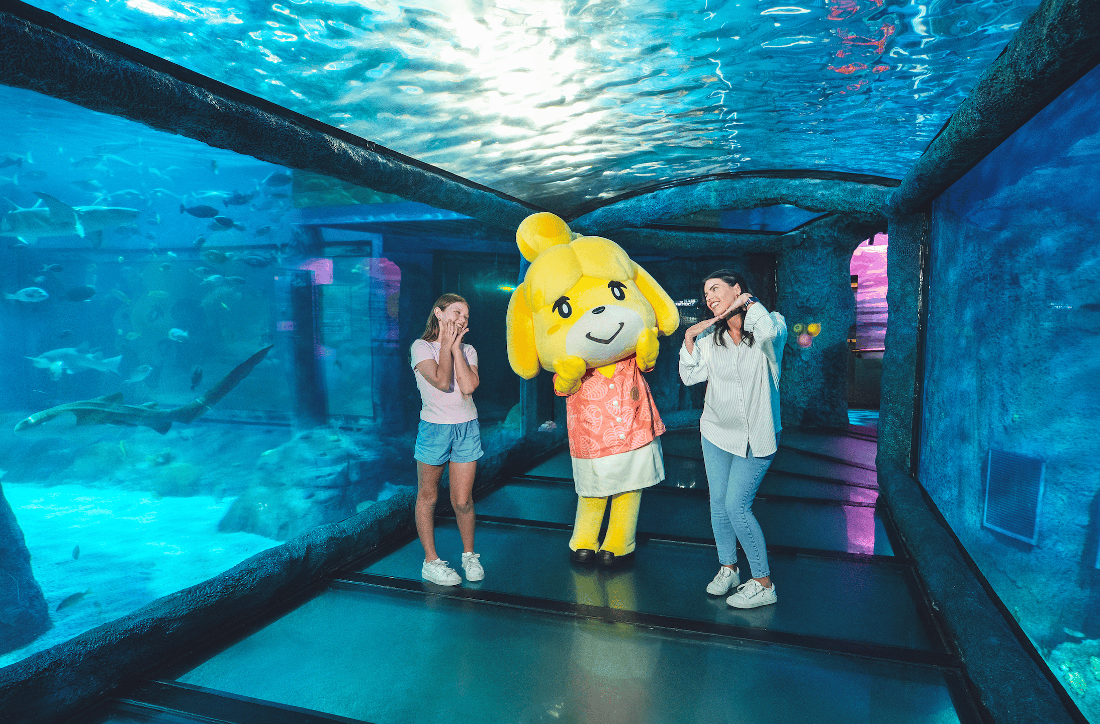 Two people pose with an Isabelle mascot costume from Animal Crossing inside an underwater aquarium tunnel, with a shark and fish swimming past the glass overhead and to the sides.