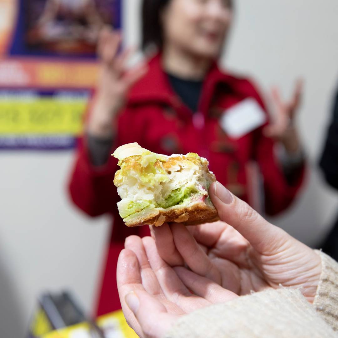 A hand holds a half-eaten sandwich, with Joyce Watts talking animatedly in the blurred background.