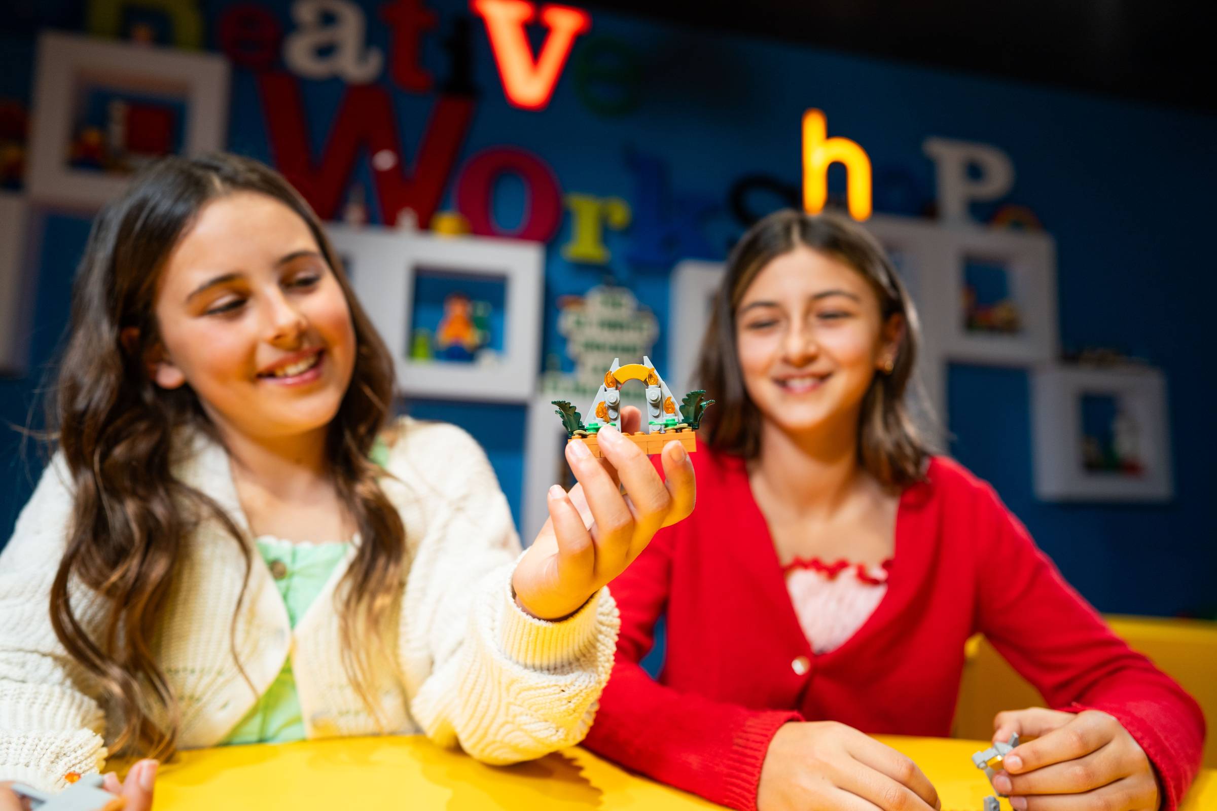 Two smiling girls sit at a yellow table in a LEGO workshop, one holding up a small brick-built jungle gate with orange flames and green leaves.