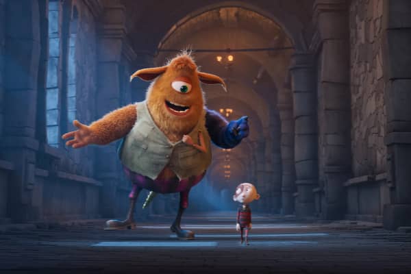An animated scene from Stitch Head showing a large, friendly one-eyed monster in a vest gesturing expressively beside a small, wide-eyed stitched boy in a gothic castle corridor.