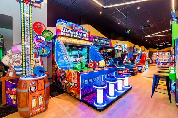 Inside an arcade venue, rows of colourful arcade machines line a corridor under blue and pink ceiling lighting. In the foreground, a barrel-shaped "Wheel of Fun" prize game sits beside a tall ticket-redemption tower, with illuminated stools lined up in front of dinosaur and racing-themed arcade games.