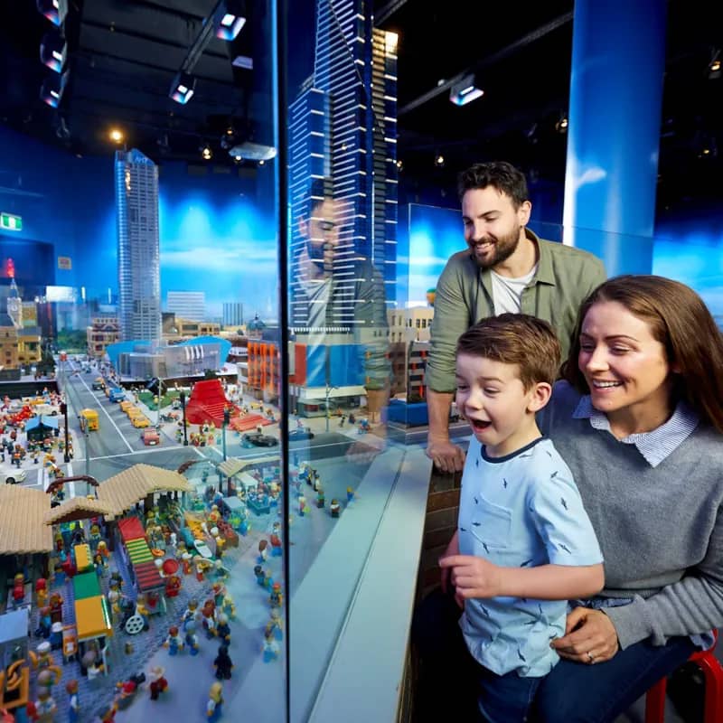 A family looks through glass at a detailed LEGO Miniland cityscape of Melbourne, lit up in blue, with a young boy's face lit up in delight as two grown-ups look on beside him.