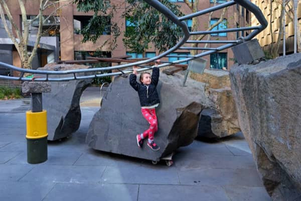 A child in a black jacket and pink leggings climbs across a large granite boulder on wheels at Rocks on Wheels playground in Southbank, gripping an overhead steel rail, with apartment buildings in the background.