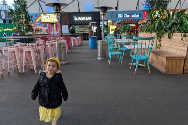 A young girl in a beanie and jacket walks through Grazeland's outdoor dining area, past colourful market stalls including The Burgie Bar and El Gaucho