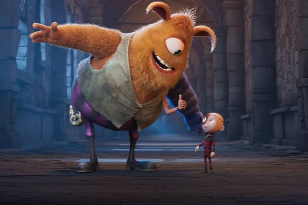 A still from the animated film Stitch Head, featuring a large, friendly-looking monster with orange fur, mismatched legs, and a patched outfit, leaning toward a small boy in a striped jumper. Set in a dark, castle-like corridor.