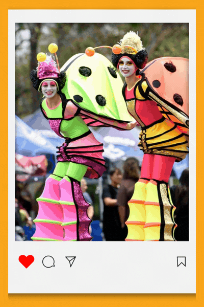 An animated social media style post in a white frame with an orange border and heart, comment, share and save icons below. It cycles through three photos from the festival:  Two performers on stilts in bright pink, green and yellow ladybird costumes with big spotted wings. A woman dressed as a fairy, in a purple hat and wings, reading aloud from a large book. A toddler in a pink jacket and beanie, held by an adult, looking at a performer in a shiny mirrored silver costume.
