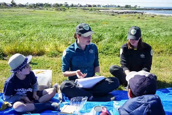 Two rangers in green uniforms sit on a picnic rug with two young children at a grassy coastal reserve.
