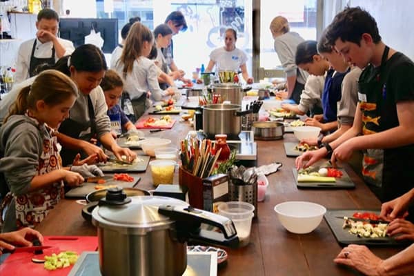 Young and older kids stand around a long table cooking and preparing various foods in a cooking class