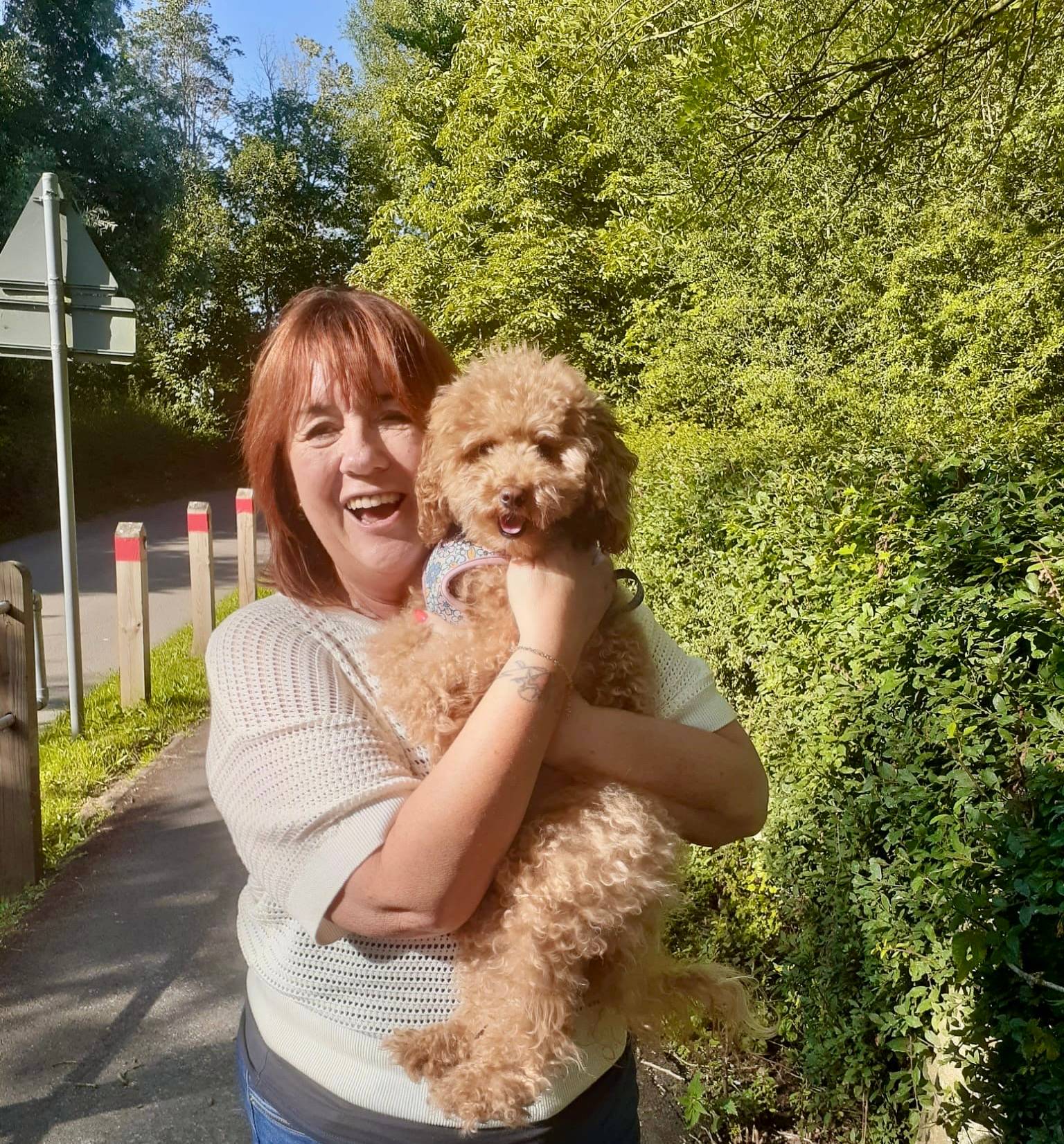 MamaJ with red hair holding blond toy poodle by green trees along a road