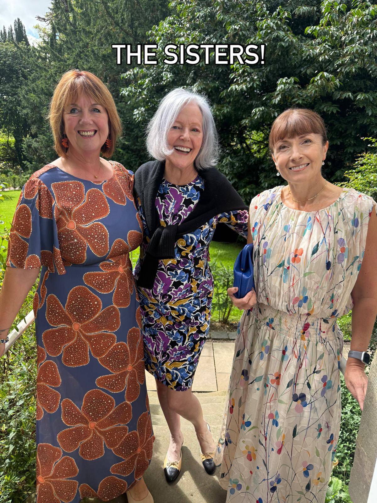 three women dressed up standing on a porch with green trees in the background