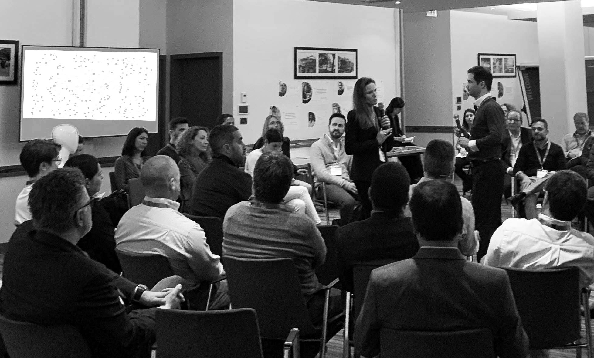 A group of people sit in chairs in a circle in a meeting room. In the center, the author and another member of the Adaptive Teams Network hold a microphone. They are speaking. In the background, there is a screen showing a network graph. On the walls, there are several pictures.