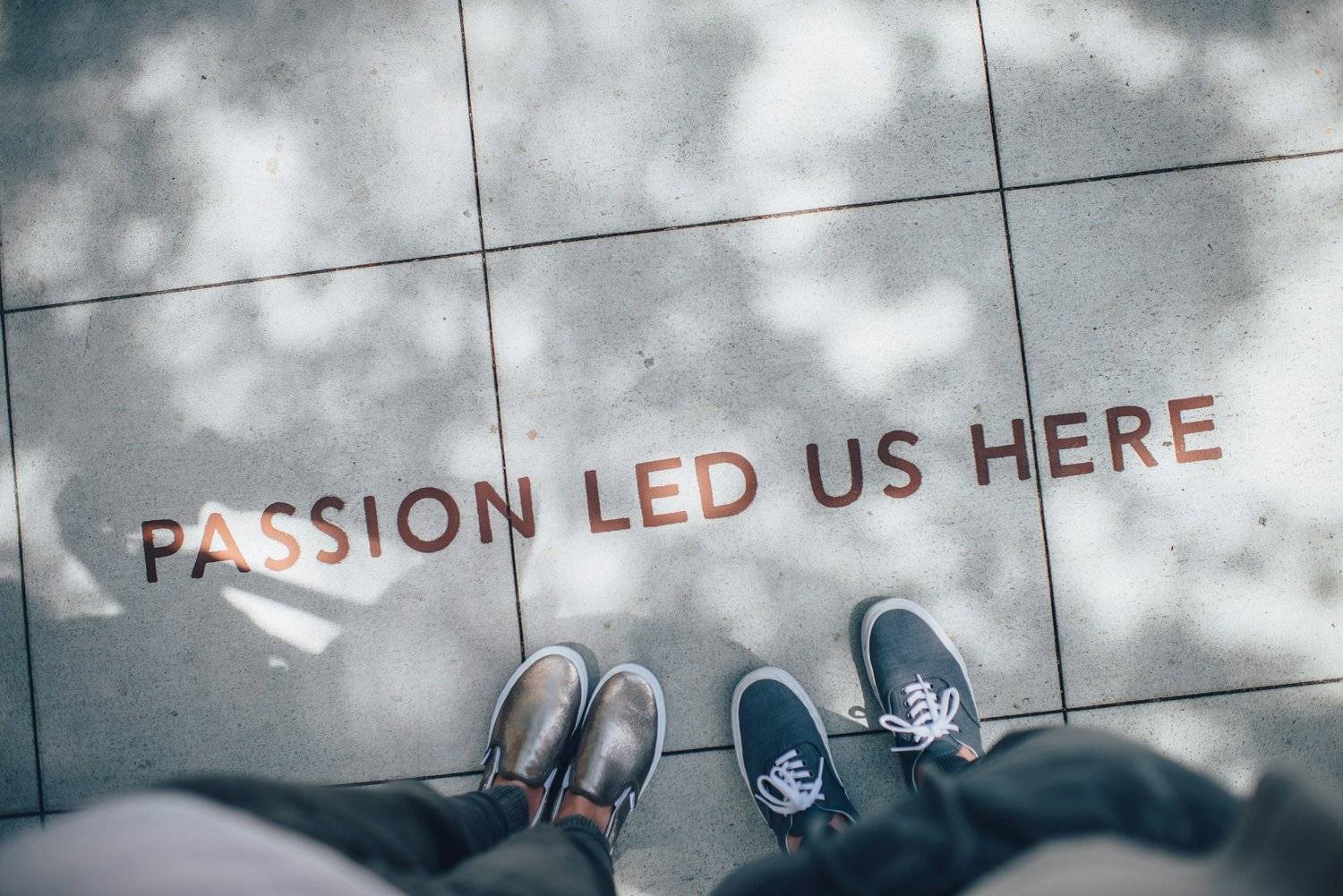 Looking down on two pairs of feet standing on a concrete sidewalk with the letters of the words "Passion Led Us Here" stenciled on the concrete. 