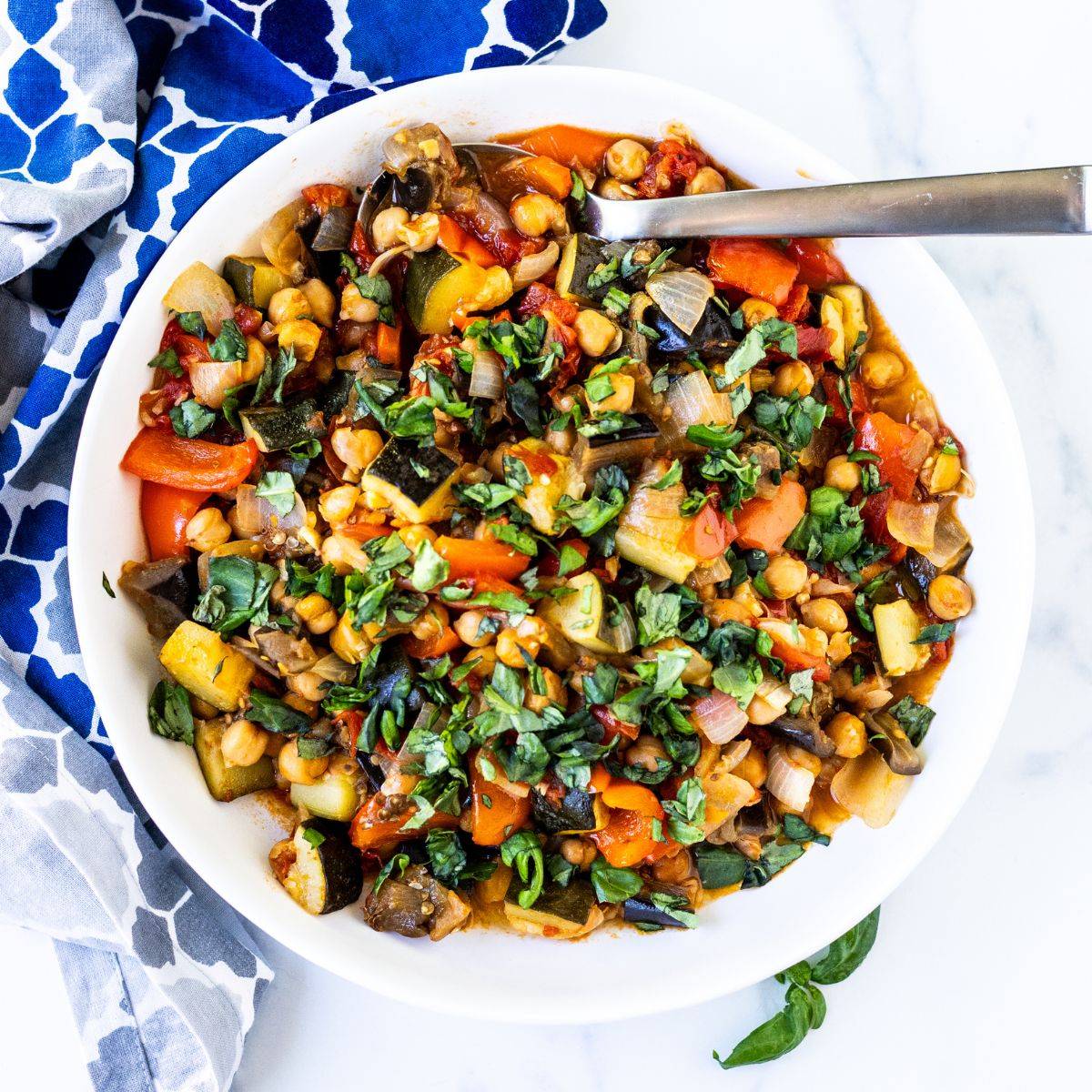Spoon in a serving bowl of roasted chickpea ratatouille garnished with fresh basil.