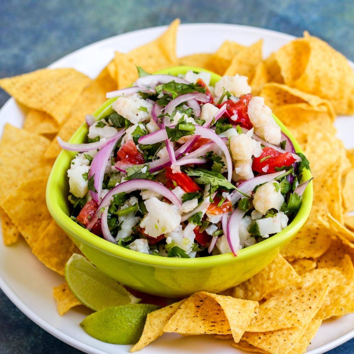 Bowl of cauliflower ceviche with red onions, tomatoes, and cilantro on a platter of tortilla chips.