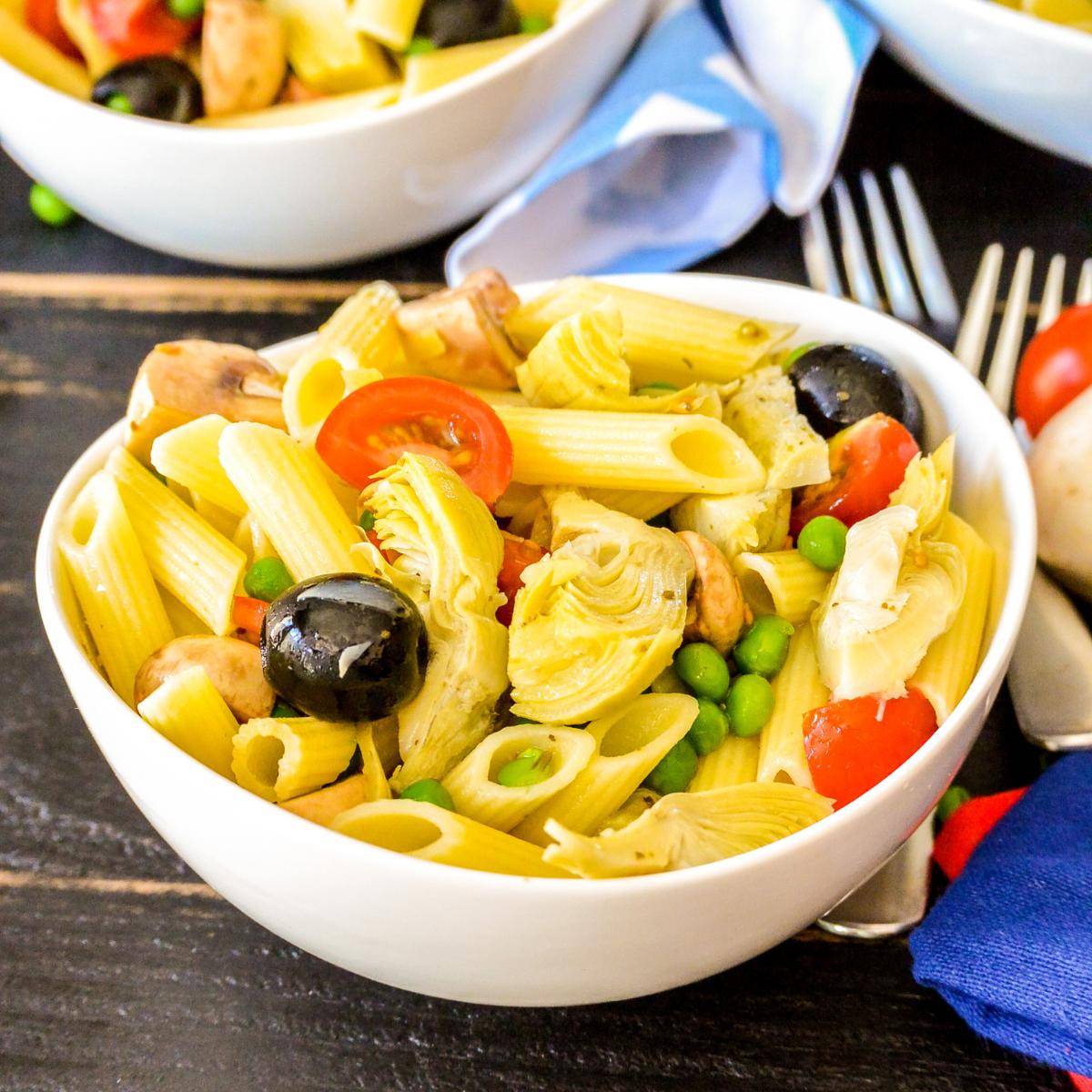 Bowl of penne pasta salad with artichoke hearts, peas, tomatoes, mushrooms, and olives.