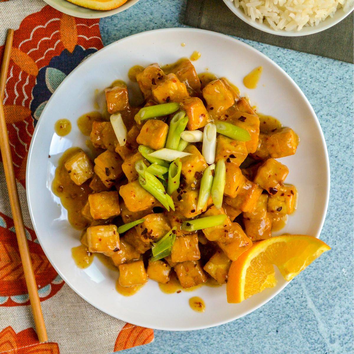 Plate of orange tofu garnished with green onions and a slice of orange.