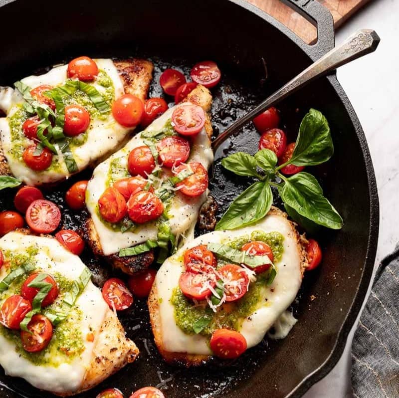 grilled chicken in a skillet covererd with mozzarella, cherry tomatoes, and pesto
