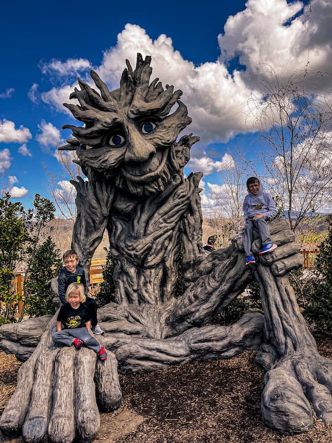 Three children sitting and climbing on a large whimsical tree-like statue with a smiling face and outstretched hands at Anakeesta in Gatlinburg, Tennessee, under a bright blue sky with scattered clouds.