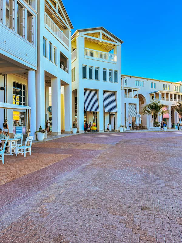 Colorful shops and restaurants with striped awnings line the town square in Seaside, Florida, with people strolling and outdoor seating on a sunny day.