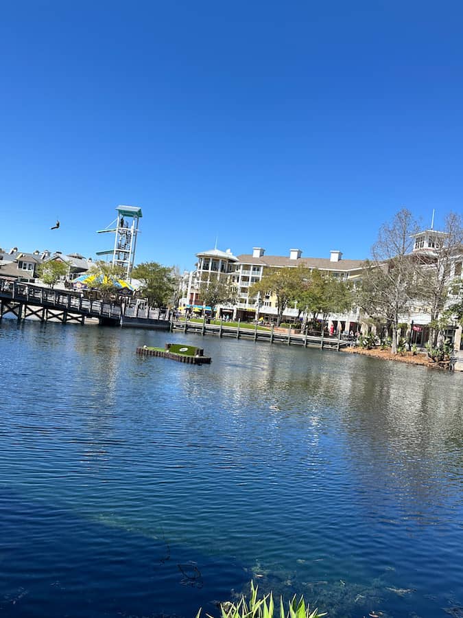 Village of Baytowne Wharf small lake with someone ziplining in the background and the large buildings with shops and restaurants in the background. 