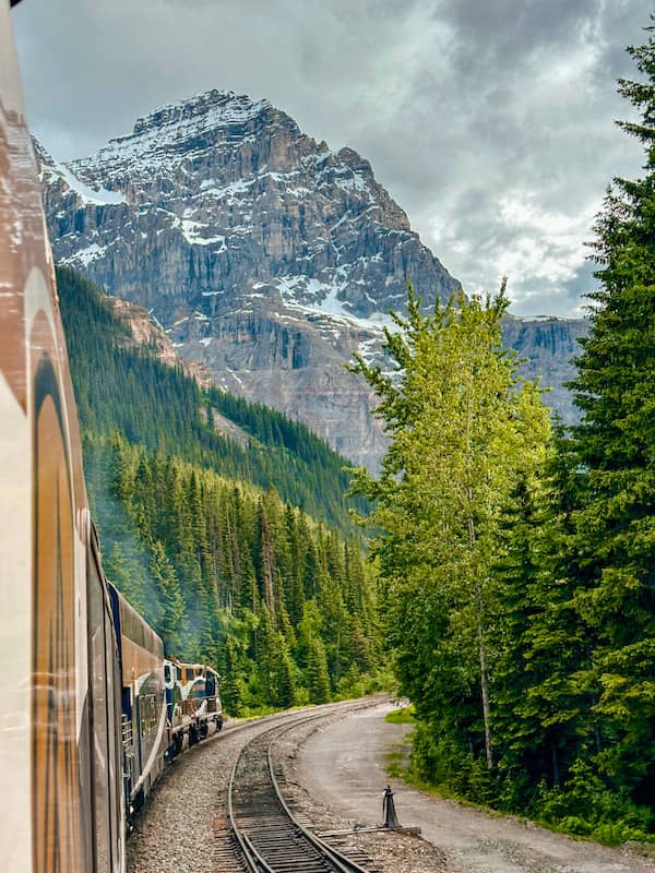 Rocky Mountaineer train rounding a curve through evergreen forest toward a snow-dusted mountain in the Canadian Rockies under cloudy skies.