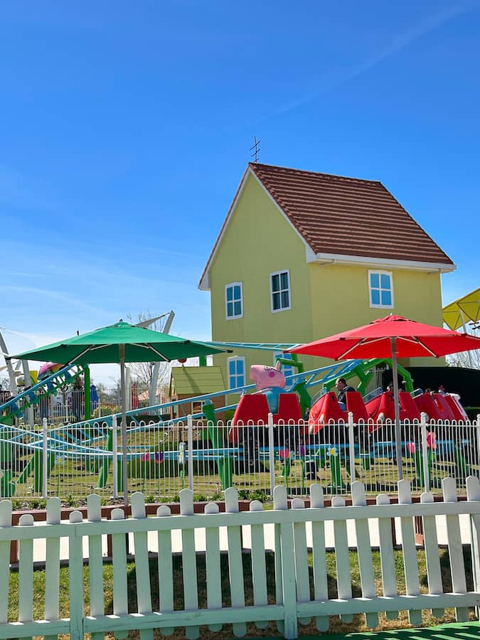 Red roller coaster with daddy Pig in the front car and Peppa's yellow house in the background at the Peppa Pig theme park.