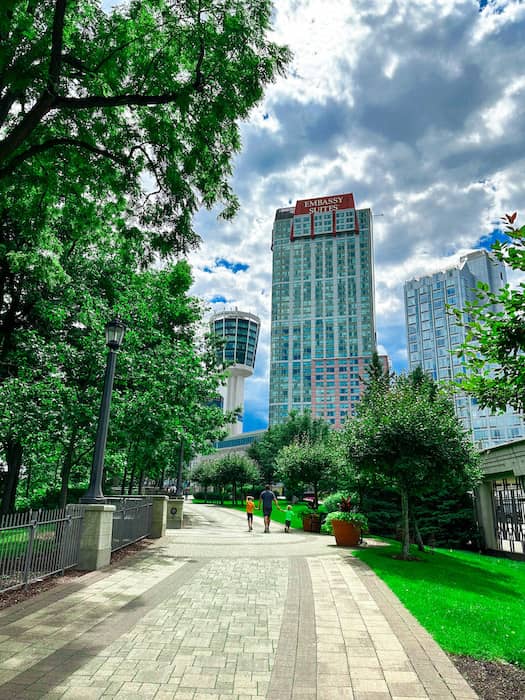 People walking down the brick walkway surrounded by many green trees and tall buildings in the background including the Embassy Suites Hotel.