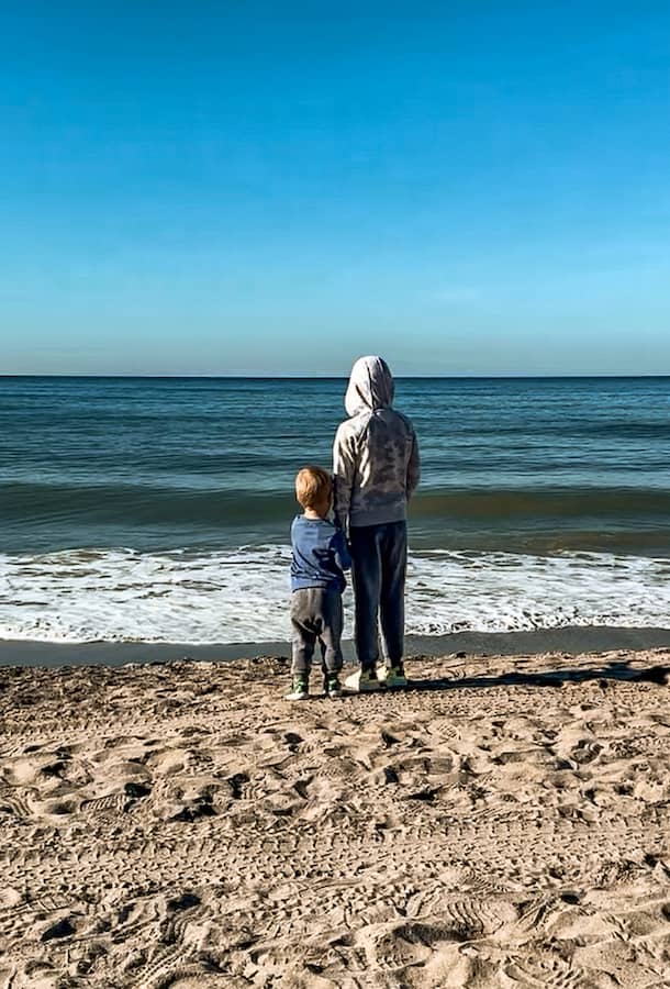 Two young boys stand side by side on a sandy beach in California, looking out at the waves of the ocean on a clear blue sky morning.