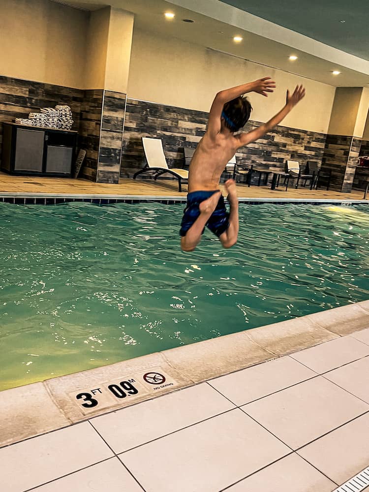 Boy jumping into an indoor hotel pool with his arms in the air and legs bent behind him.