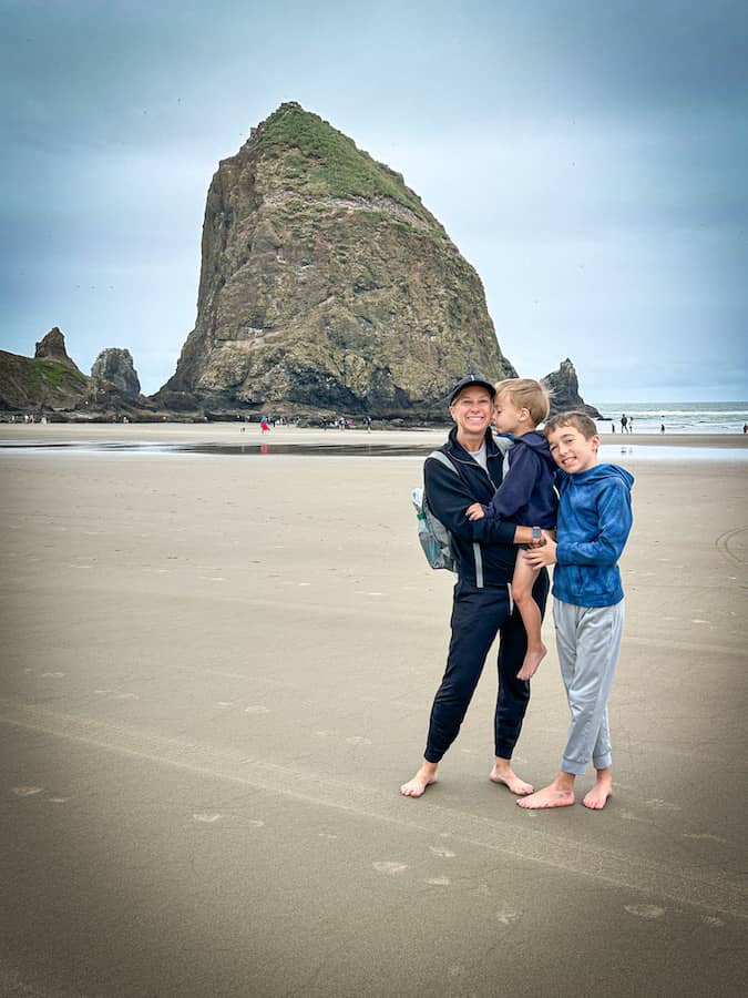 Mom holding a young boy while another young boy is leaning on them in front of Haystack Rock on Cannon Beach, Oregon.