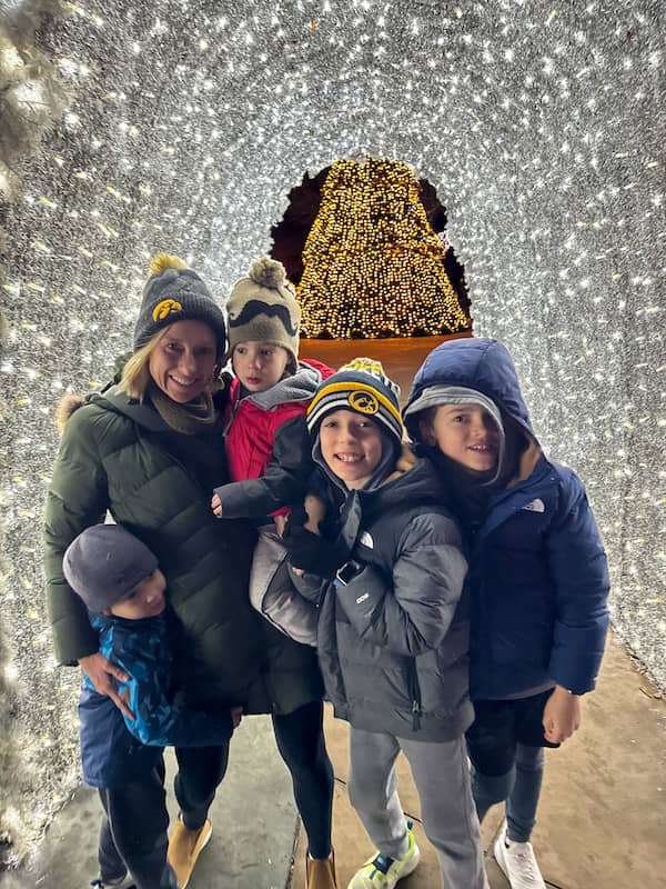 Mom and four boys bundled in winter coats beneath a tunnel of sparkling white holiday lights, with a golden Christmas tree glowing behind them.