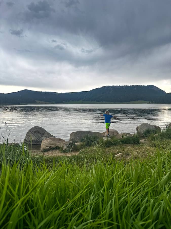 Young boy throwing rocks into Meadowlark Lake in Wyoming with mountains and storm clouds in the distance.