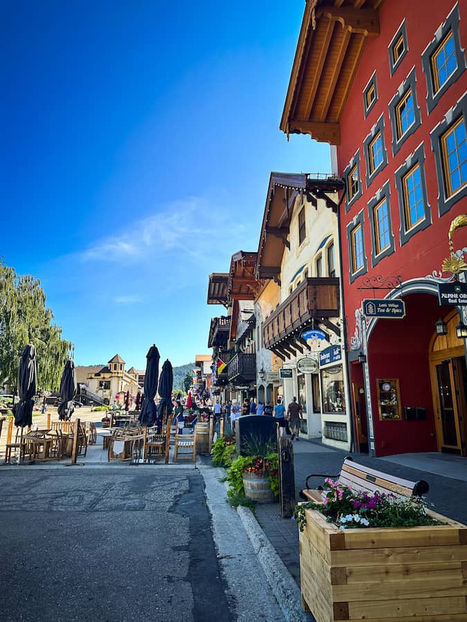 Colorful Bavarian-style buildings line a charming street in downtown Leavenworth, Washington, with outdoor café tables, flower planters, and a bright blue sky overhead.