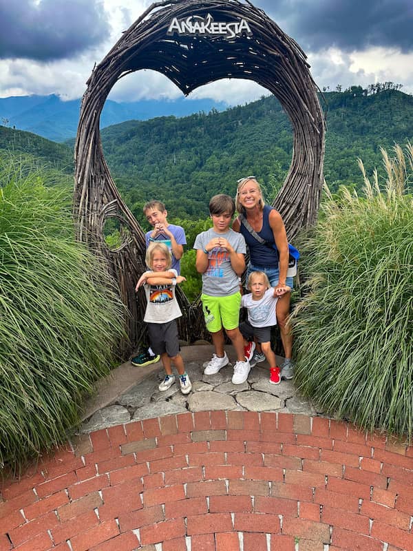 Mom with four young boys posing for a picture with the mountains in the background at Anakeesta in Gatlinburg. The mom is holding one boy's arms as he looks unhappy. The other boys are making heart with their hands or crossing their arms revealing the lit