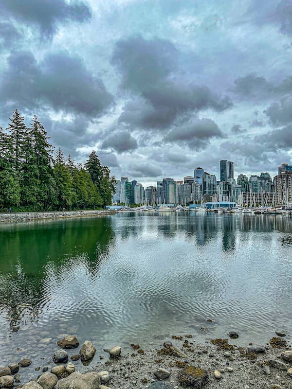 View of Vancouver skyline from Stanley Park shoreline with calm water, boats in the marina, and cloudy skies overhead