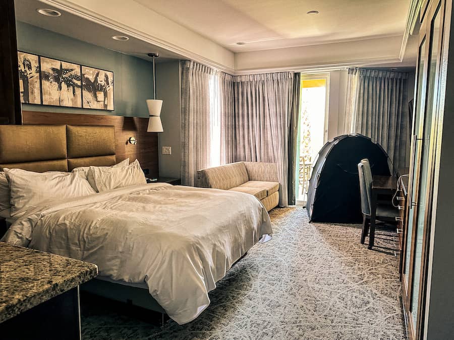 pacious hotel room with a king bed, sofa, desk, and a small pop-up sleep tent for kids set up near the balcony—perfect setup for a family stay.