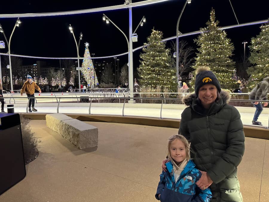 Mom with her son wearing winter coats and hats posing in front of the ice rink and lighted trees in Heartland of America Park in Omaha, Nebraska.