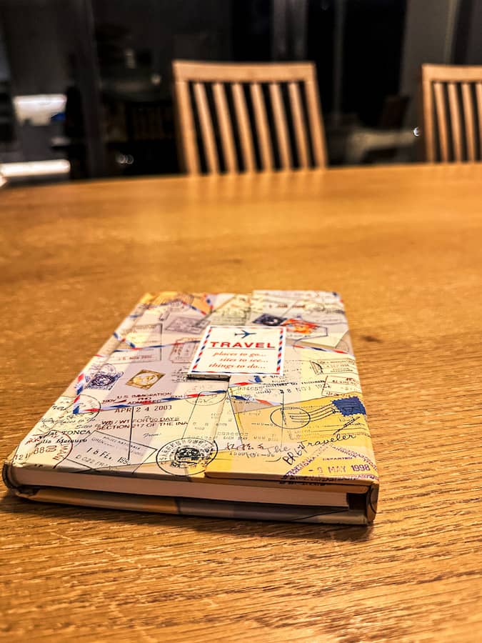 Travel journal with maps and passport stamp illustrations resting on a dining room table while planning an upcoming family road trip.