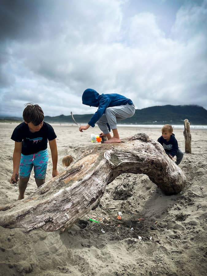 Three young boys play and climb on a large piece of driftwood on the sandy beach in Seaside, Oregon, under cloudy skies with grassy dunes and coastal hills in the background. 