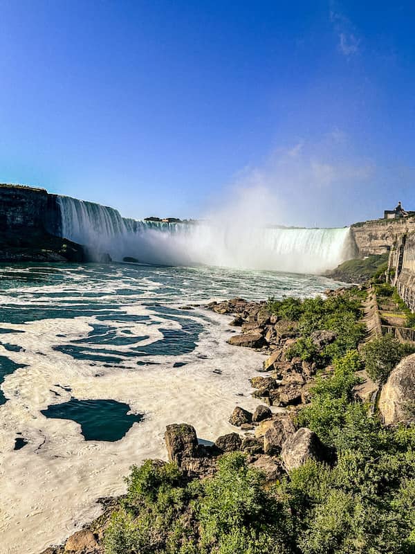 View of Horseshoe Falls in Niagara Falls, Ontario, with mist rising above the waterfall and the Niagara River below on a sunny day.