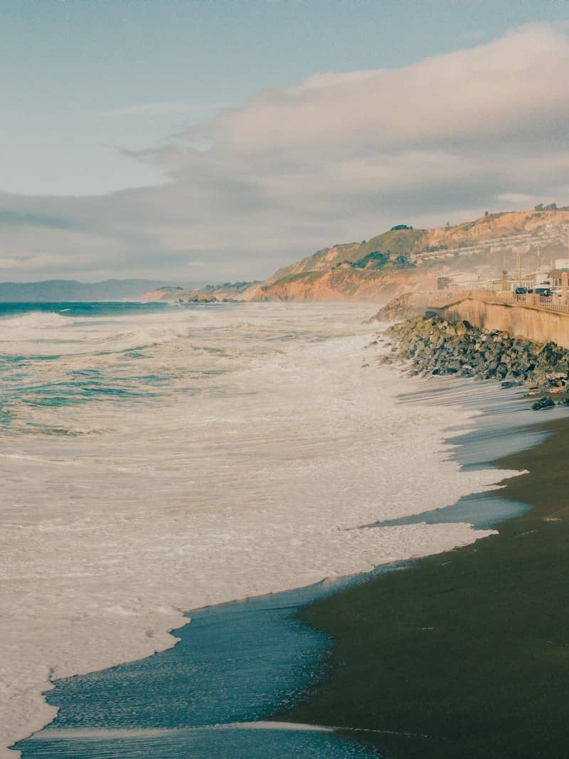 Waves crashing onto a sandy beach below a coastal cliff with houses