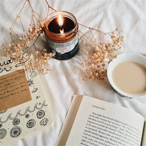 A cozy flat lay featuring an open journal with doodles, an open book, a lit soy candle in a glass jar, delicate dried flowers, and a cup of coffee or tea, all arranged on a soft white blanket—evoking a calm, reflective, and self-care-focused atmosphere.
