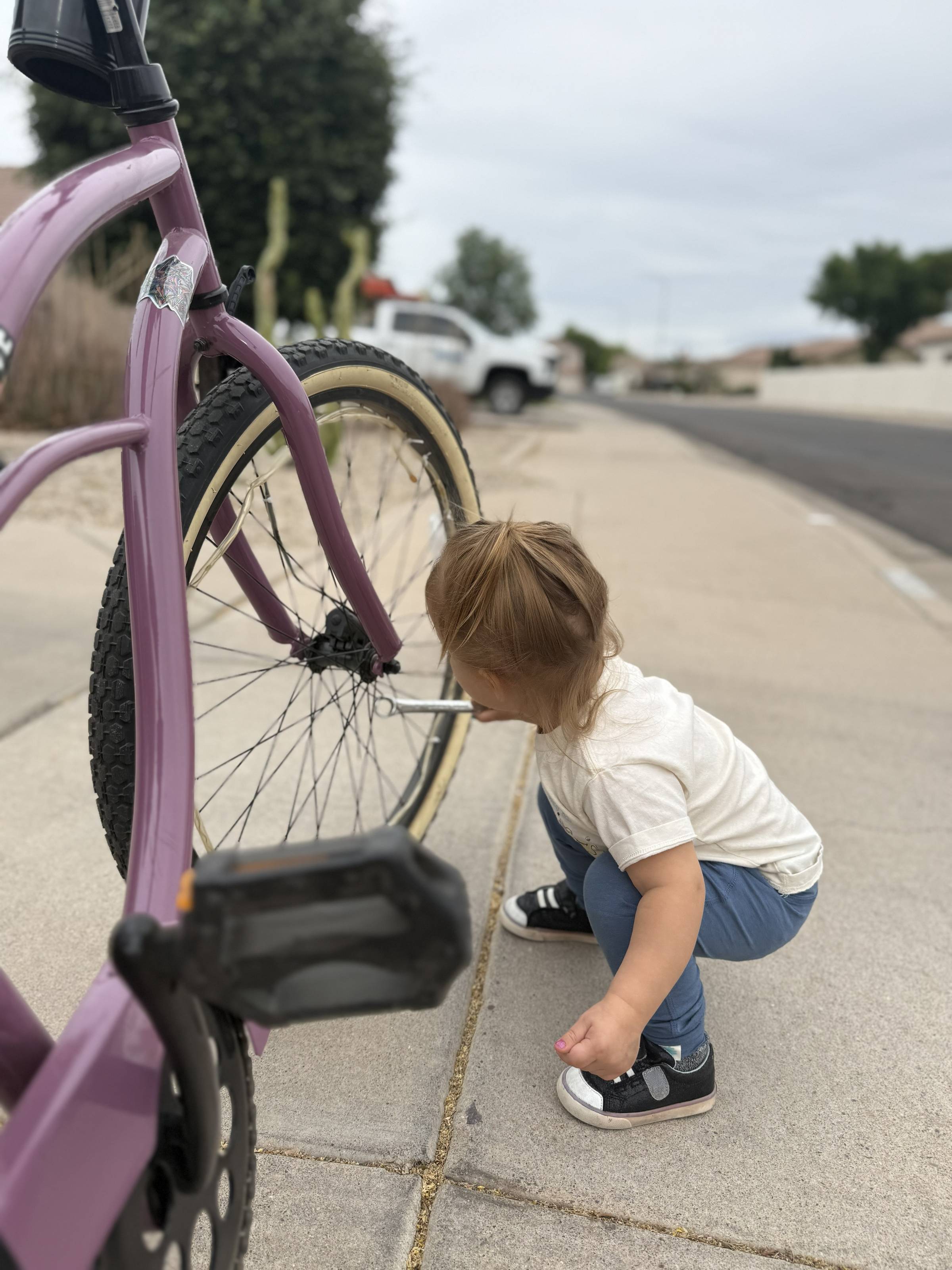 Toddler girl crouching on a sidewalk, closely examining the wheel of a purple bicycle with curiosity and focus.