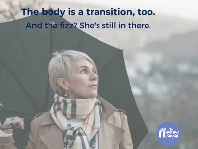 A midlife woman with short silver hair, wearing a tan coat and plaid scarf, holds an open umbrella on an overcast day and gazes quietly into the distance. Text overlay reads: "The body is a transition, too. And the fizz? She's still in there." — Find Your