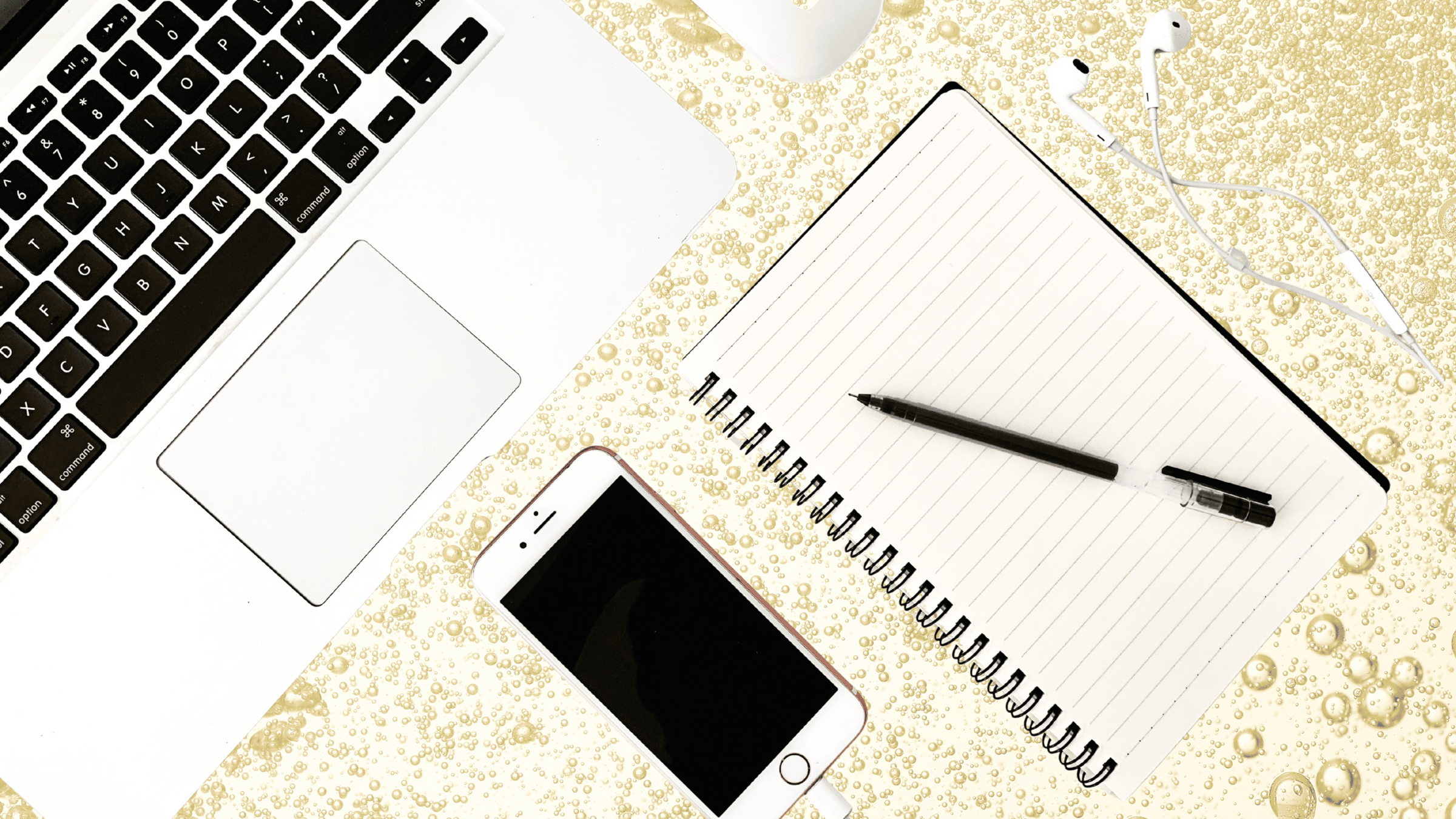 A flat lay of a workspace with a silver laptop, spiral notebook with pen, white earphones, and a smartphone on a bubbly champagne-colored background.