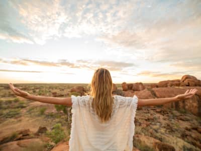 A woman with blonde hair stands on rocky terrain at sunrise, arms outstretched toward the horizon. She wears a light, flowing white top, symbolizing freedom, renewal, and openness to possibility.