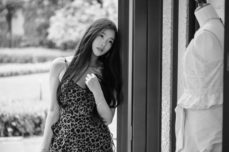 Woman in patterned dress leaning against doorframe, looking at mannequin.