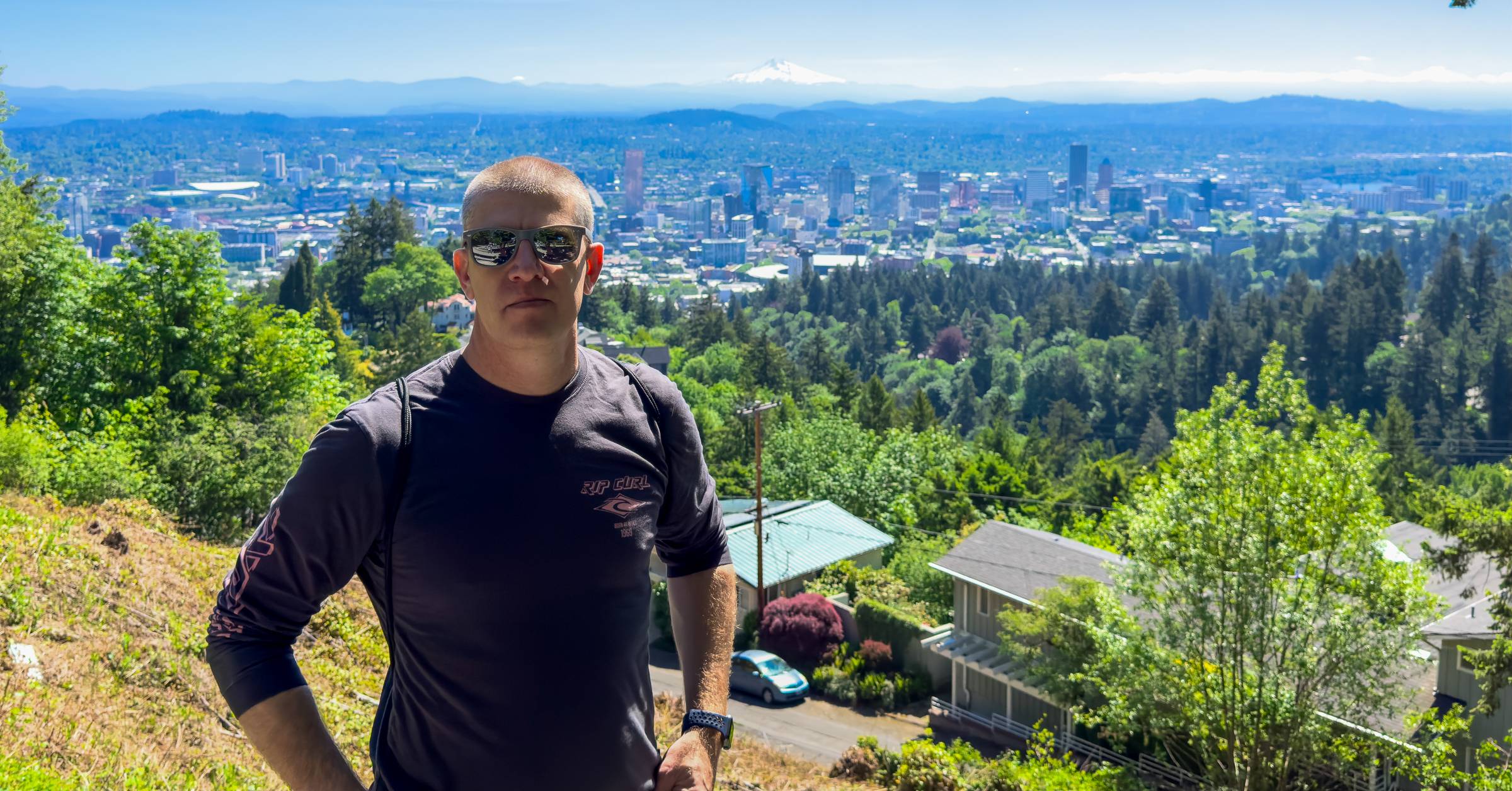 Man wearing sunglasses and a black long sleeve shirt stands on a hillside overlooking a city skyline surrounded by trees. Snow capped mountain is visible in the distance under a clear blue sky, adding depth to the scenic urban view.