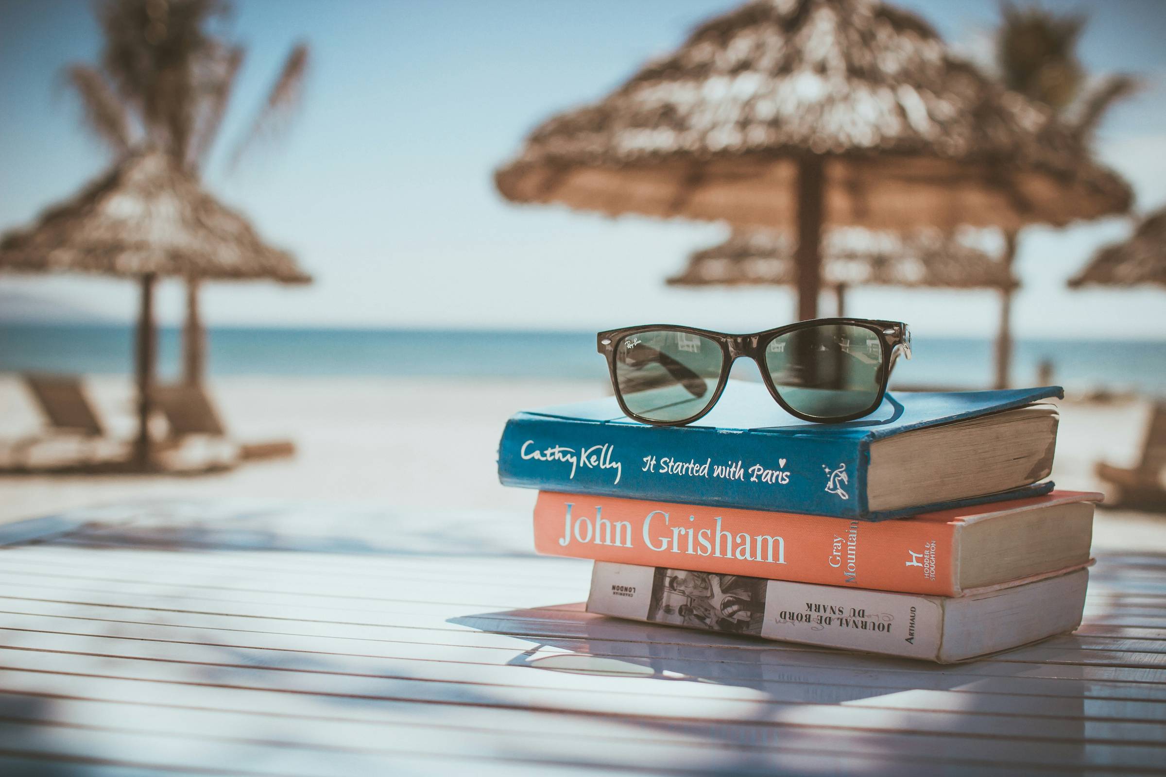 A pile of 3 books and a pair of sunglasses on a table with a beach, straw umbrellas, and ocean in the background. 